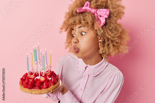 People and celebration. Indoor sideway photo of young pretty African american female having party standing in centre isolated on pink background holding cake with strawberries blowing candles