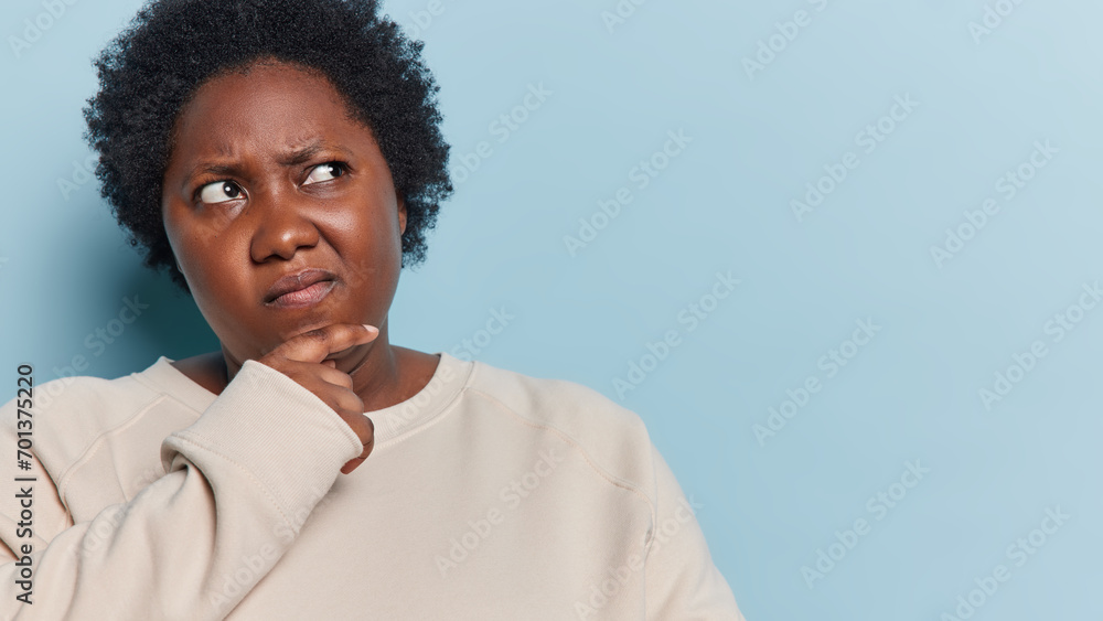 Obraz premium Studio shot of pensive dark skinned plump African woman keeps hand on chin concentrated aside being deep in thoughts wears casual jumper isolated over blue background with copy space for your text