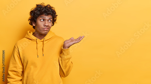 Confused hesitant Hindu man shrugs shoulder keeps palm raised up dressed in casual sweatshirt doesnt know why happened like this isolated over yellow background copy space for your promotion photo