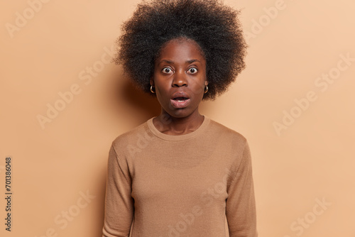 Waist up shot of shocked African woman with dark bushy hair has scared horrified expression dressed in casual clothing holds breath from amazement isolated over brown background. Omg concept