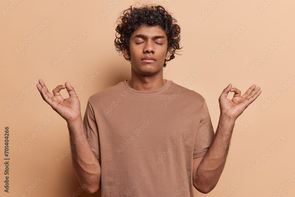 People and peace. Studio shot of young calm relaxed Hindu man standing in centre isolated on beige background with closed eyes meditating to relax and calm down after working day dressed in t shirt People and peace. Studio shot of young calm relaxed Hindu man standing in centre isolated on beige background with closed eyes meditating to relax and calm down after working day dressed in t shirt