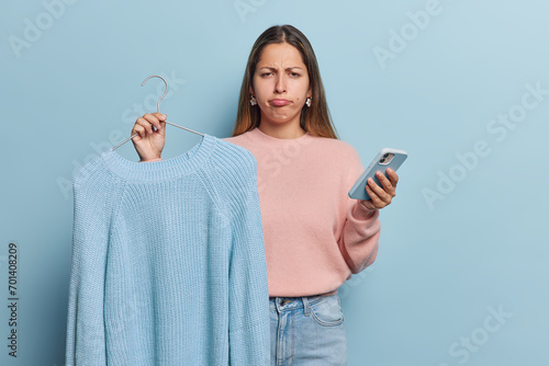 Photo of discontent European woman holds jumper on hanger uses mobile phone for surfing internet purses lips being dissatisfied with her purchase isolated over blue background. Shopping concept