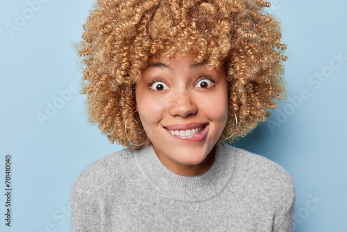 Portrait of surprised curly haired woman bites lips and stares with widely opened eye has curious expression wears casual grey jumper isolated over blue background. Human facial expressions concept photo