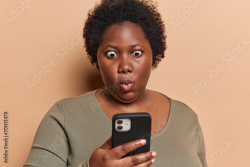Photo of stupefied dark skinned woman checkes mailbox reads disturbing message received fascinating offer holds smartphone feels impressed wears casual jumper isolated over brown background. photo
