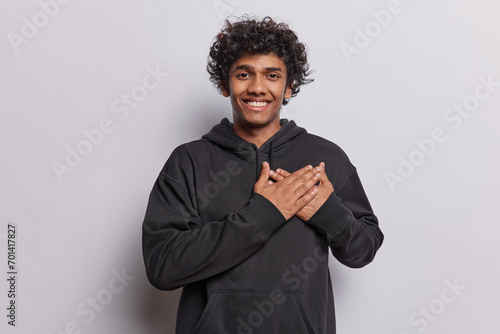 Waist up shot of happy Hindu man with curly hair keeps hands on heart expresses gratitude feels flattered and thankful smiles gladfully wears black sweatshirt isolated over white background. photo