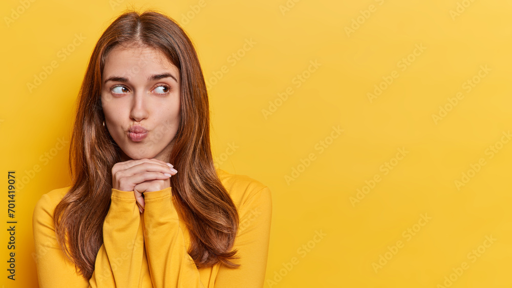 Pensive long haired woman keeps lips folded hands under chin concentrated aside with thoughtful expression dressed in casual jumper isolated over yellow background copy space for your promotion Pensive long haired woman keeps lips folded hands under chin concentrated aside with thoughtful expression dressed in casual jumper isolated over yellow background copy space for your promotion