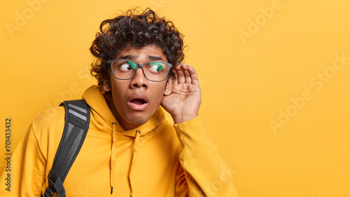 Horizontal shot of curly haired Hindu man with curly hair keeps hand near ear tries to overhear something eavesdropping for fresh gossips dressed in casual sweatshirt isolated over yellow background