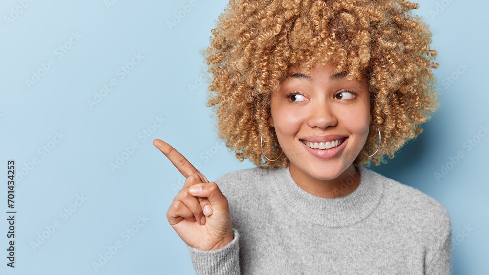 Studio shot of curly haired woman dressed in casual grey jumper points index finger on blank space for your advertisement isolated over blue background. People advertisement and promotion concept - obrazy, fototapety, plakaty 