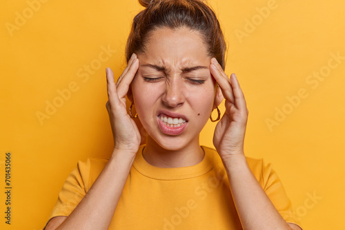 Exasperate young woman suffers from terrible headache keeps hands on temples clenches teeth keeps eyes closed wears casual t shirt isolated over vivid yellow background. Health problems concept photo