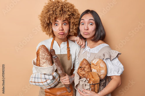 Indoor shot of pair of young surprised Asian and African american women standing in centre close to each other on brown background holding homemade hot tasty loaves of bread after cooking in aprons