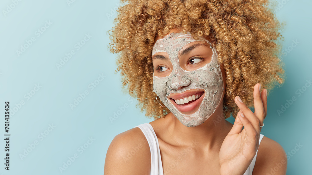 Beautiful cheerful curly haired woman applies facial clay mask for skin treatment focused aside smile gently undergoes beauty procedures after shower isolated over blue background copy space Beautiful cheerful curly haired woman applies facial clay mask for skin treatment focused aside smile gently undergoes beauty procedures after shower isolated over blue background copy space