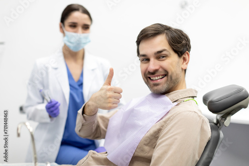 Happy male patient showing thumb up and smiling at camera with female doctor dentist sitting on background. Healthy teeth concept