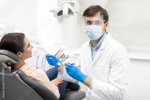 Male dentist smiling and working in dentist office, repairing teeth of young woman, dentist looking at camera wearing white uniform, mask and dentistry glasses