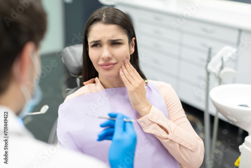 Young woman in dental chair suffering from acute tooth pain, touching cheek and looking at doctor
