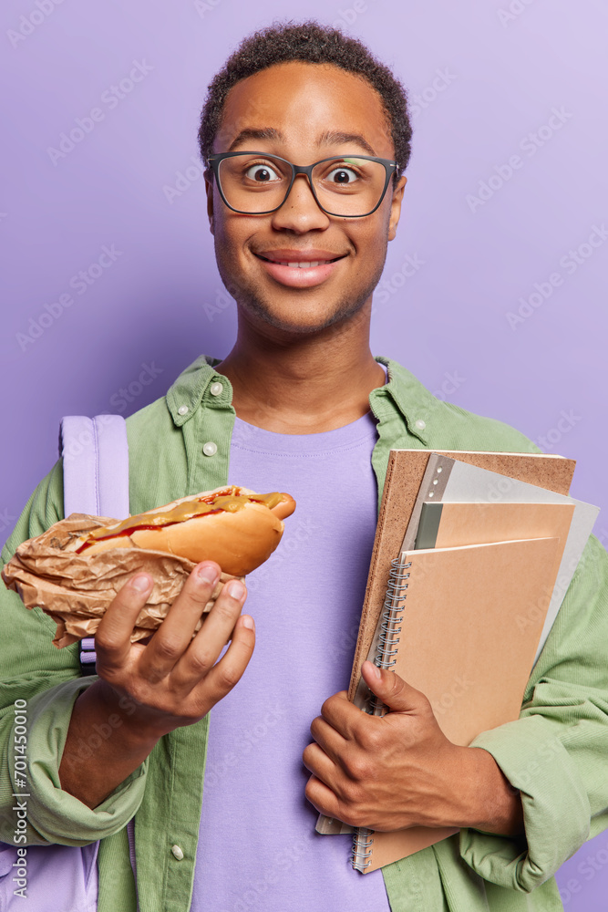 Vertical image of surprised dark skinned curly man holds hot dog and notepads eats during break wears optical eyeglasses and shirt carries backpack on shoulder isolated over purple background. Vertical image of surprised dark skinned curly man holds hot dog and notepads eats during break wears optical eyeglasses and shirt carries backpack on shoulder isolated over purple background.