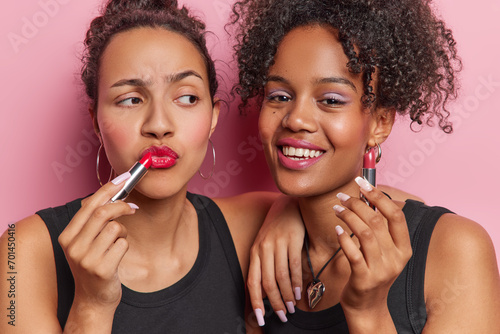 Beauty concept. Two women apply cosmetic hold lipstick and smile happily prepare for party dressed in black t shirts isolated over pink background. Female friends put on makeup before dating