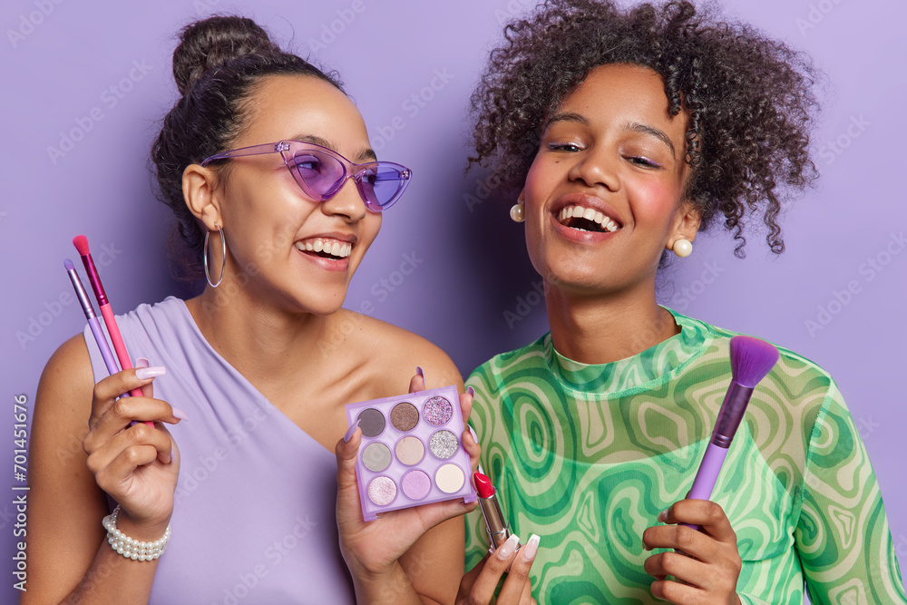 Overjoyed women prepare for dating laugh happily and pose with cosmetic products apply eyeshadow and lipstick dressed in fashionable clothes isolated over purple background. Beauty treatments concept - obrazy, fototapety, plakaty 
