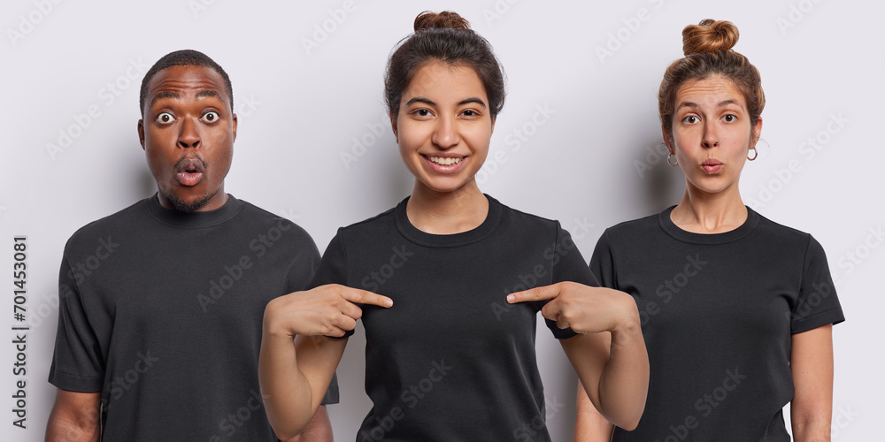 Smiling Iranian woman points at mock up space of casual t shirt being in good mood stands in centre shocked African man and amazed female model stand against white background. Collage image. - obrazy, fototapety, plakaty 