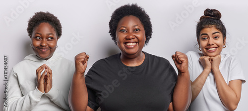 Horizontal shot of African woman steeples fingers looks aside happy overweight dark skinned female model cheerful Iranian girl keeps hands under chin stand next to each other isolated on white wall