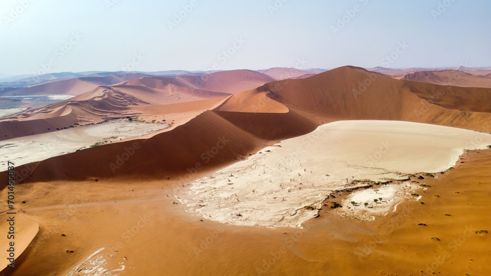 Naklejka premium Deadvlei, Namibia - August 24, 2022: The stark contrast of Deadvlei's white pan, dotted with the skeletal remains of trees, is framed by the towering rust-red dunes under the clear blue sky.