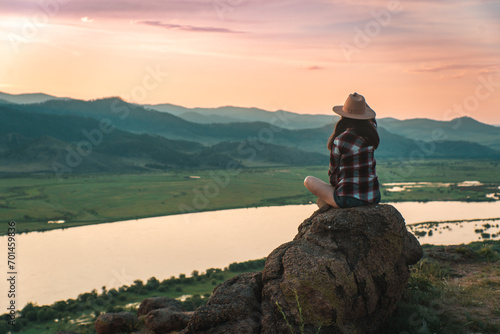 hatted woman in checkered shirt on the top of the mountain in summer against the sunset over the river.  the persin right sided of  the frame photo