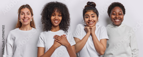 Horizontal image of cheerful mixed race young women look gladfully at camera press hands to heart feel thankful smile gladfully express positive emotions dressed in casual basic white clothes. photo
