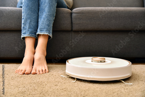 Modern robot vacuum cleaner near sofa with woman legs in room