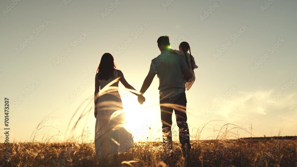 Silhouette of couple with daughter sitting in father arms walking in field in evening. Silhouette of married couple walking in field with daughter in arms. Father and mother with child walk at sunset - obrazy, fototapety, plakaty 