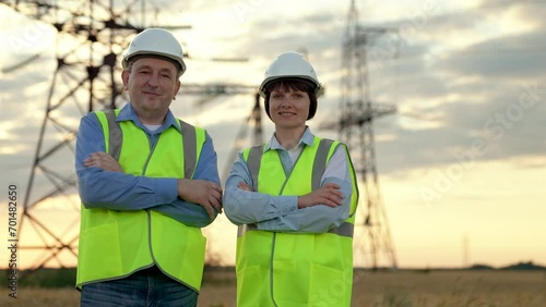 Cheerful electricians colleagues stand crossing arms against electric power transmission lines in sunset field. Engineers team with crossed arms rest after work at power distribution substation photo