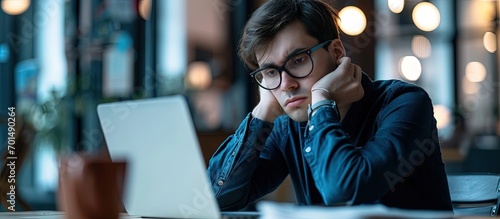 Young unhappy man office worker feeling bored at work looking at laptop with demotivated face expression while sitting at workplace in office distracted male worker feeling tired of monotonous photo