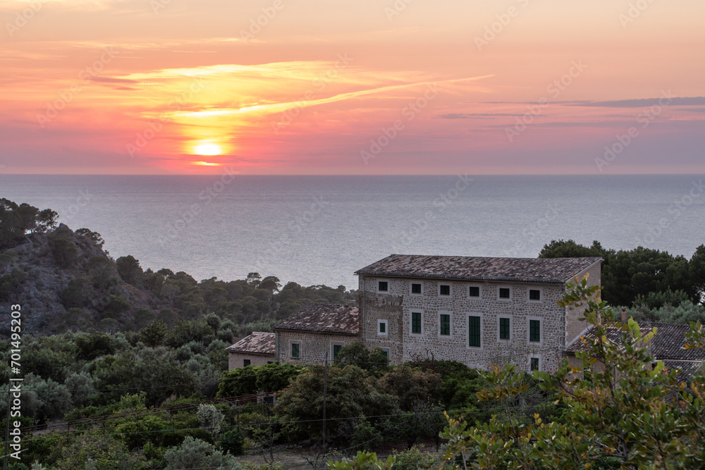 A Mediterranean-style stone building surrounded by nature and with a dramatic sunset over the Mediterranean Sea on the island of Majorca, Spain