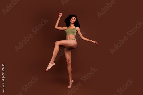 No filter full length photo of cheerful positive sporty girl with thin slim body raise leg isolated on brown color background