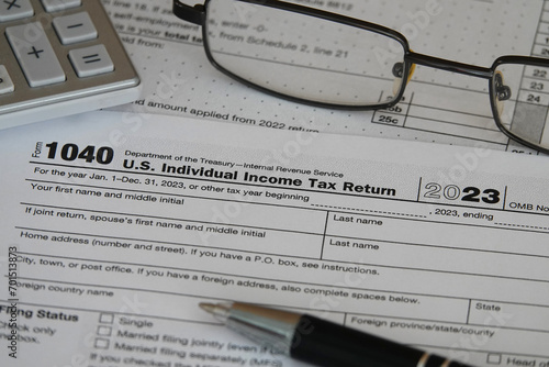 An IRS 1040 tax year 2023 form is shown in 2024, along with an ink pen, calculator, and glasses. The Internal Revenue Service tax filing deadline in 2024 is scheduled for April 15.