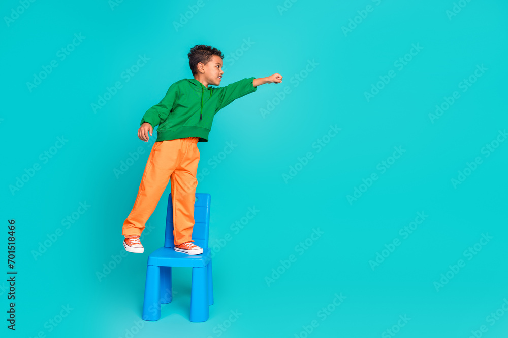 Full length photo of crazy nice stylish boy standing on chair raised hand looking empty space isolated on cyan color background Full length photo of crazy nice stylish boy standing on chair raised hand looking empty space isolated on cyan color background