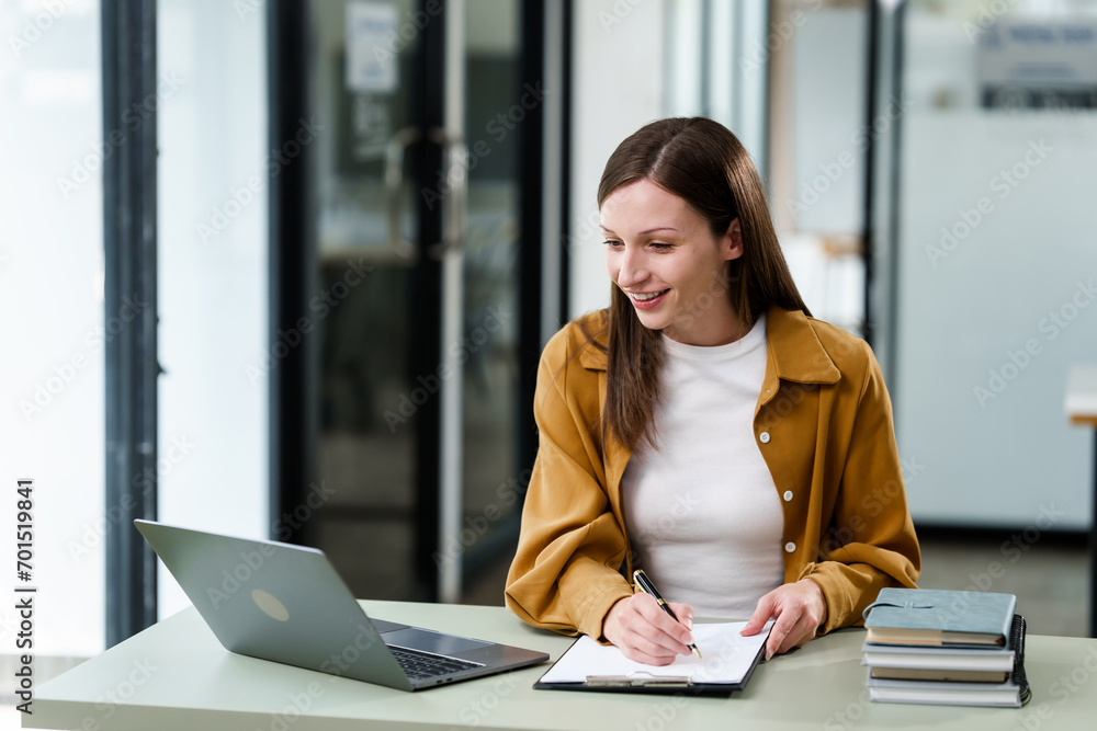 Caucasian university woman working on a project, with a laptop, clipboard in hand, and books on the table. Caucasian university woman working on a project, with a laptop, clipboard in hand, and books on the table.