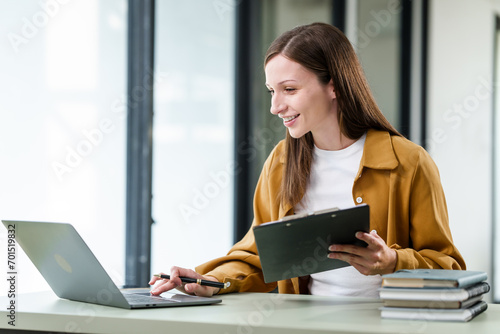 Caucasian university woman working on a project, with a laptop, clipboard in hand, and books on the table.