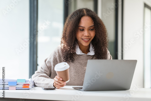 African American university student with a coffee cup, engaged in her studies or working on her MBA on a laptop, with books stacked beside her.