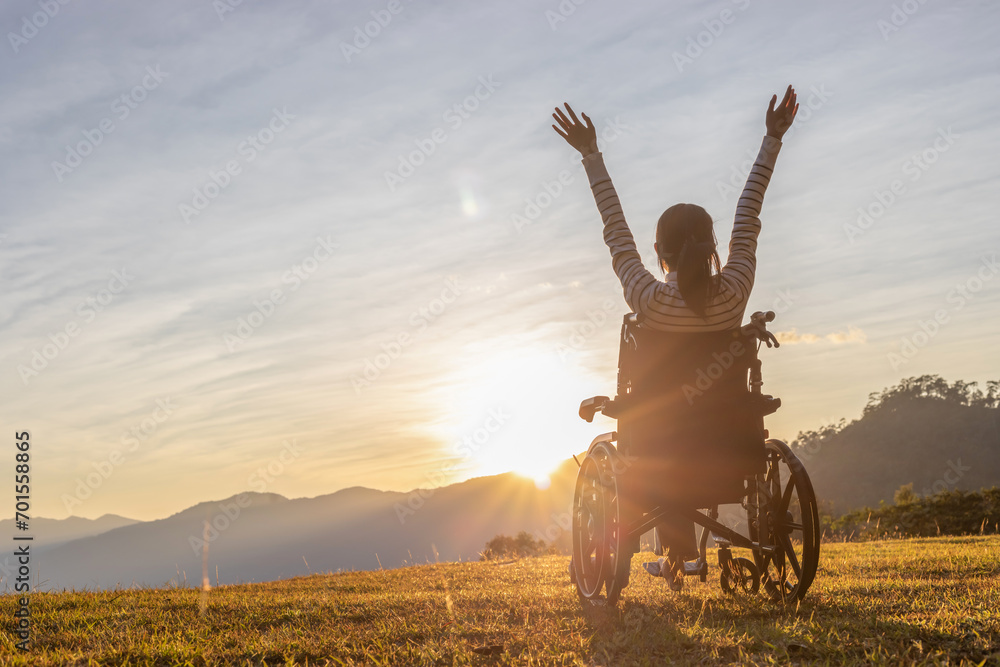 Person sitting on a wheelchair enjoying with raised hands on the mountain at the sunset time. - obrazy, fototapety, plakaty 