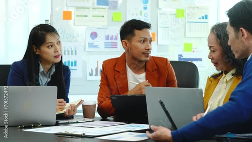 Asian business manager is leading a meeting, engaging with trainees in a bright room with data charts, indicating a monthly review or training session.