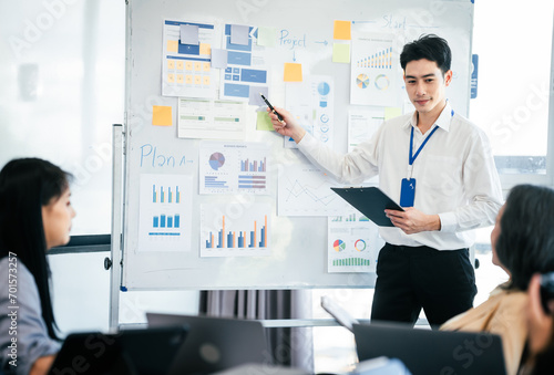 mature Asian business manager is leading a meeting, engaging with trainees in a bright room with data charts, indicating a monthly review or training session.