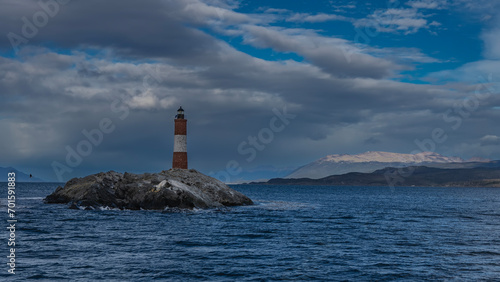 The old famous lighthouse Les Eclaireurs at the edge of the world. The red-and-white striped tower stands on a small rocky island in the Beagle Channel. Ripples on the water. Clouds in the blue sky. 
