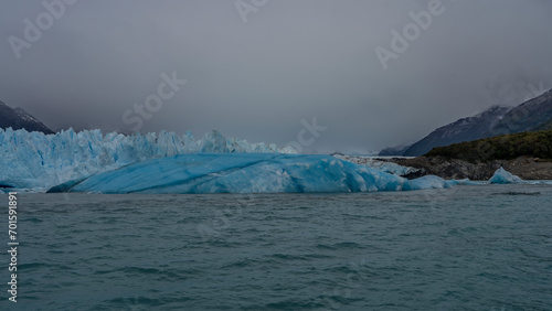 The famous Perito Moreno Glacier. A wall of blue ice against a cloudy sky and mountains. A huge melting iceberg floats in a turquoise glacial lake. El Calafate. Argentina. Los Glaciares National Park