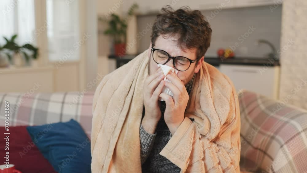 Seasonal Ailments Man with Glasses, Flu Stricken, Duvet Covered, Wiping Nose on Sofa at Home Seasonal Ailments Man with Glasses, Flu Stricken, Duvet Covered, Wiping Nose on Sofa at Home