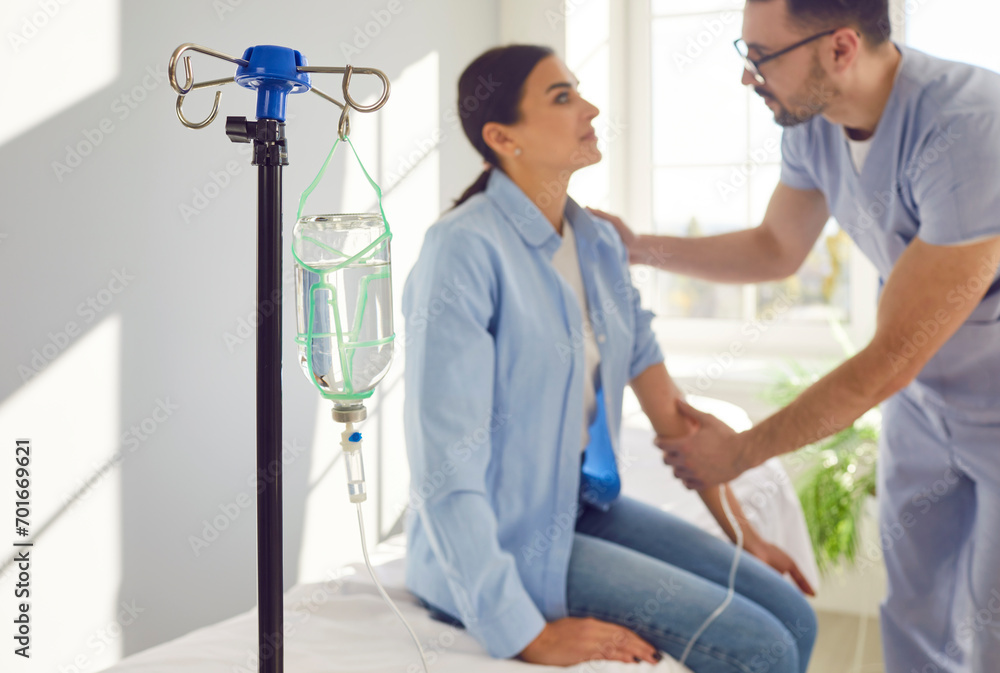 Obraz premium Male nurse sets up IV line and calms down scared female patient. Close up drip bottle with medicine in hospital ward, man doctor giving intravenous line to worried woman in background. Medical concept