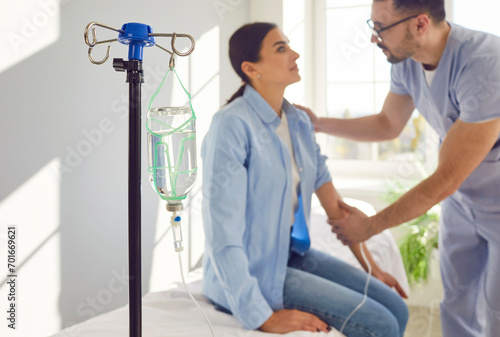 Male nurse sets up IV line and calms down scared female patient. Close up drip bottle with medicine in hospital ward  man doctor giving intravenous line to worried woman in background. Medical concept