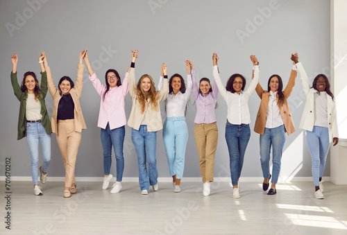 Group of funny business multiethnic women looking at camera, holding hands up and smiling on a grey wall background. Company employees happy to make a good deal. Team work concept.