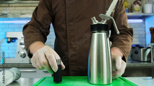 Chef in gloves changes co2 charger of whipping siphon at table closeup. Man uses modern molecular gastronomy equipment in kitchen