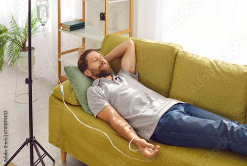 Man lies on comfortable sofa under saline drip in modern private luxury hospital. Young satisfied Caucasian man rests lying under intravenous drip. Concept of healthcare and medicine. 