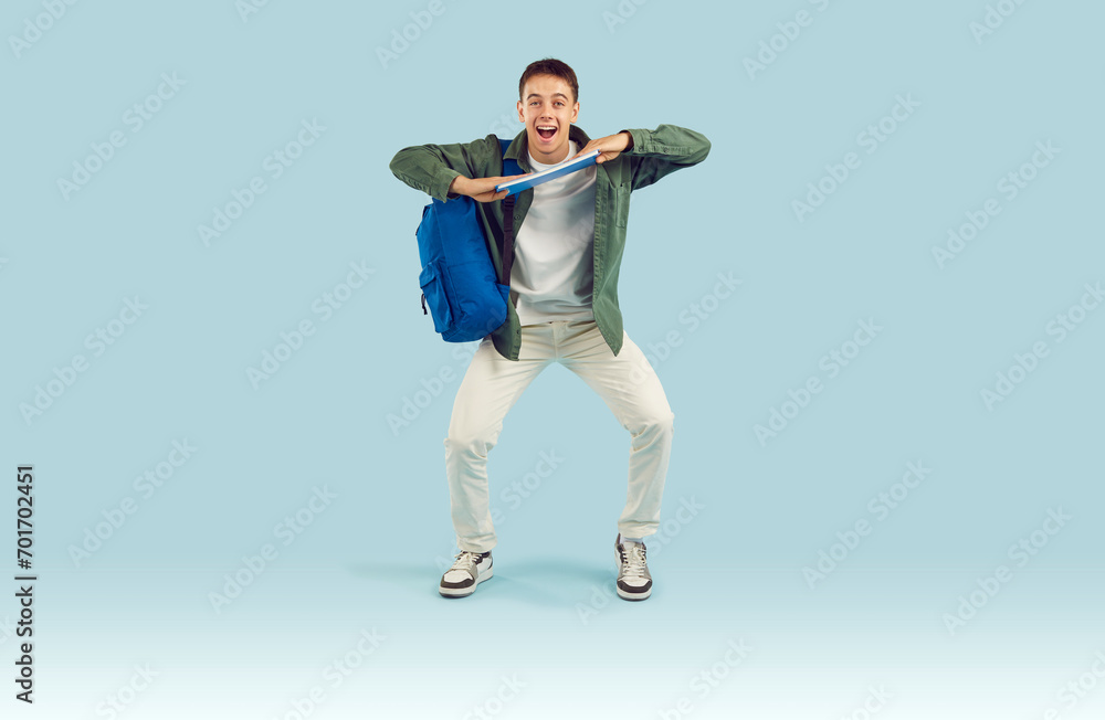 Happy adolescent male high school, college or university student having fun in studio. Full body length funny excited young man or teenage boy with book and backpack dancing on light blue background