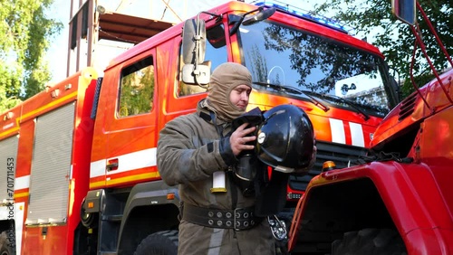 Male firefighter in uniform taking off helmet stands near a fire engine. Young fireguard removing equipment from himself after fire against background of a big red truck. Concept of saving lives photo